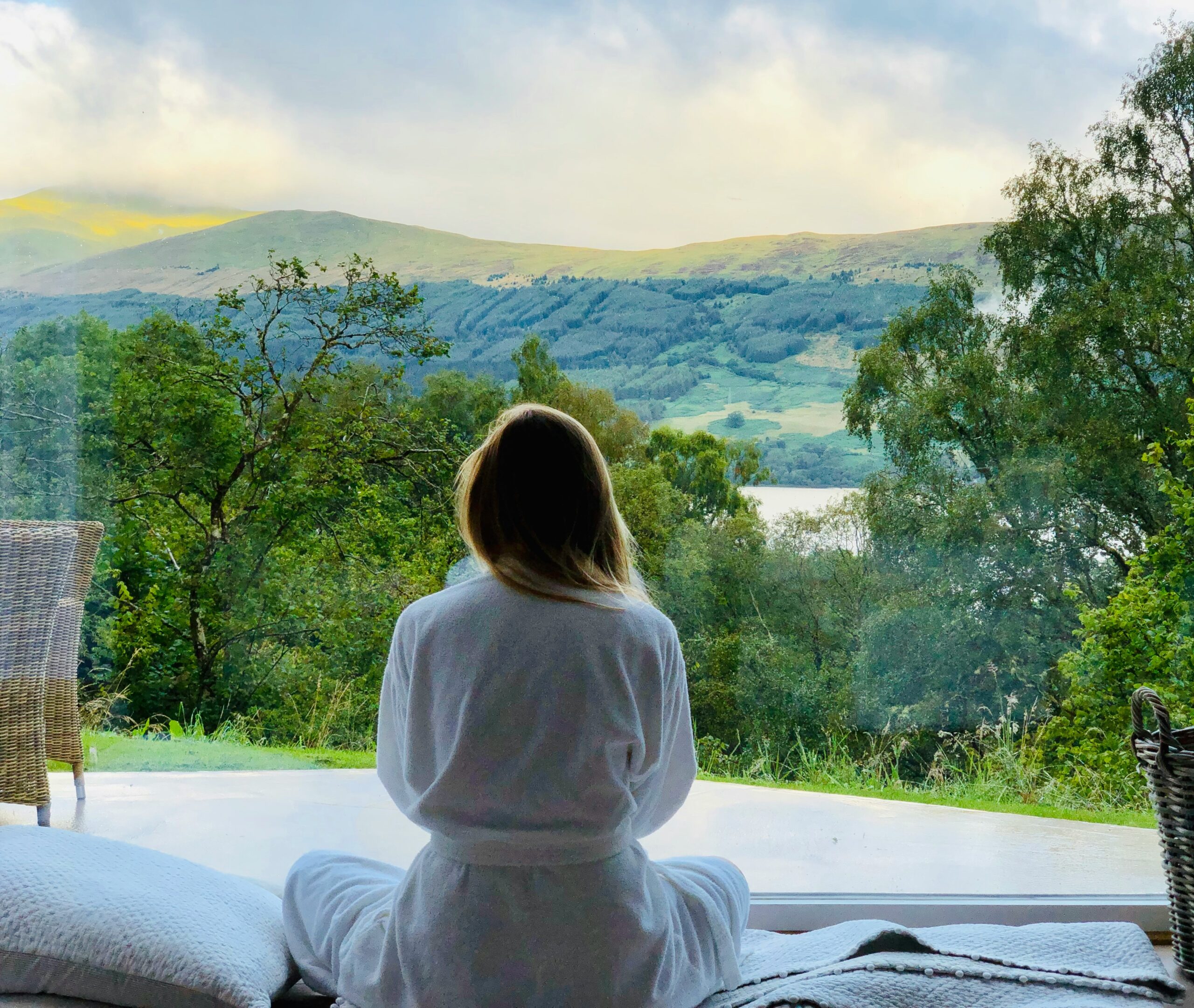 Woman in white robe sitting in front of a window looking out into the mountains.