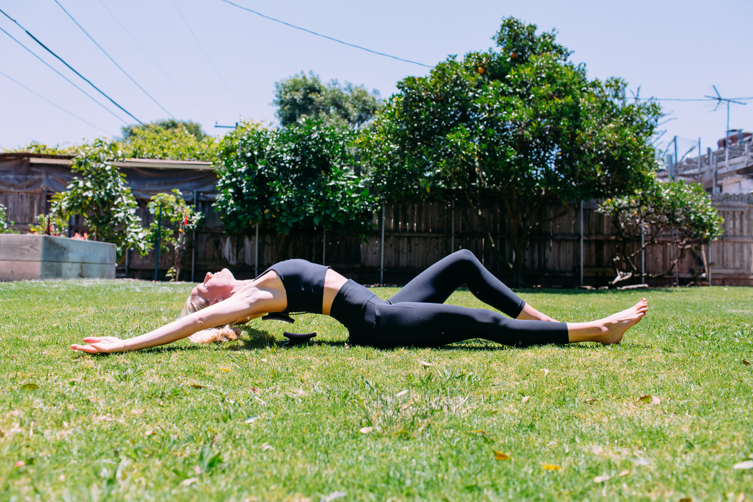 Woman stretching with a pilates ring outside on the grass with trees in the background.