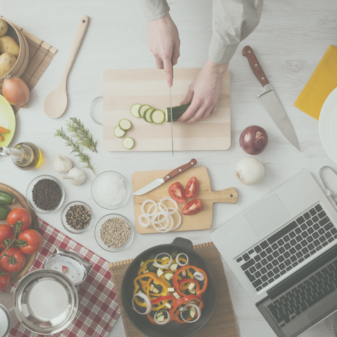 Picture of someone chopping up cucumbers with a variety of vegetables on the counter and a laptop in the corner where the recipe is.