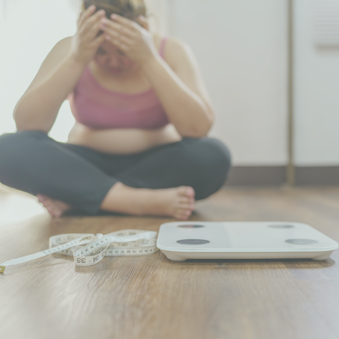 Overweight woman sitting on the floor behind a scale and tape measure with her head in her hands because she's upset.